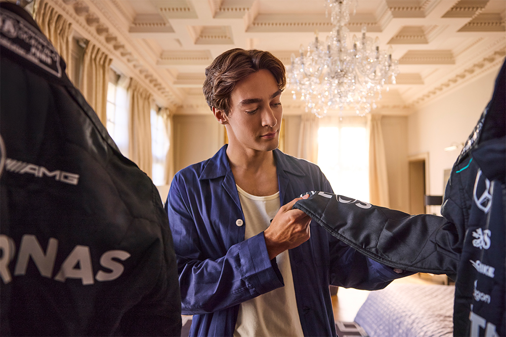 George Russell thoughtfully examines a Mercedes-AMG PETRONAS racing suit in an ornate room.