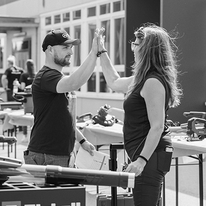 A man and a woman give each other a high five at an outdoor Einhell event with tool displays, shown in black and white.