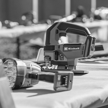 An Einhell cordless jigsaw stands on a table, with a cordless lamp in front, black and white photo.
